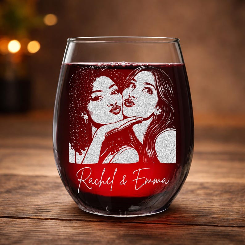 May include: Stemless wine glass filled with red liquid, featuring an etched design of two women blowing kisses. The names "Rachel & Emma" are scripted below the image. The glass sits on a wooden surface.