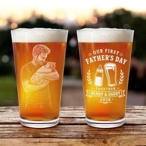 May include: Two pint glasses filled with beer, featuring personalized engravings. One glass has a father holding a baby, the other reads "Our First Father's Day Together Henry & Daddy 2026" with a bottle and beer mug.