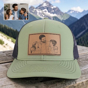 May include: A sage green trucker hat with a dark gray mesh back and a brown leather patch. The patch features a sketch of a man and two children. The hat is sitting on a wooden surface with a mountain backdrop.