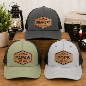 May include: Three trucker hats with mesh backs and leatherette patches. The patches read "GRANDPA", "PAPAW", and "POPS", each with "EST. 2026" and a mountain graphic. The hats are gray, olive green, and light gray.