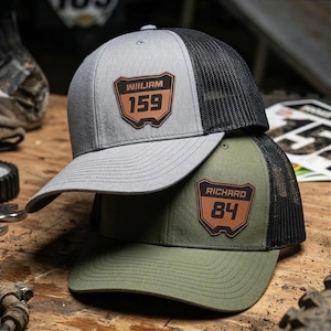 May include: Two trucker hats, one gray and one olive green, stacked on top of each other. The gray hat has a black mesh back and a brown leather patch with the name "WILLIAM" and the number "159". The olive green hat has a black mesh back and a brown leather patch with the name "RICHARD" and the number "84".