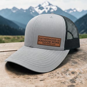 May include: Gray trucker hat with a black mesh back and a curved brim. A rectangular brown patch on the front displays the text "FATHERHOOD IS KINGDOM WORK PROVERBS 22:6". The hat is on a wooden surface with a mountain backdrop.