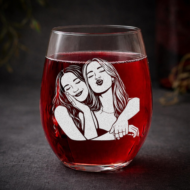 May include: Stemless wine glass filled with red liquid, featuring an etched design of two women embracing. The design is in white, with one woman resting her head on the other's shoulder.