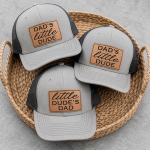 May include: Three gray trucker hats with black mesh backs and curved visors are displayed in a woven basket. Each hat features a brown leather patch with the words "DAD'S LITTLE DUDE" or "LITTLE DUDE'S DAD" in black lettering.