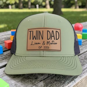 May include: A green and black trucker hat with a brown leather patch. The patch reads "TWIN DAD" above "Liam & Mateo" and "Est. 2026". The hat has a curved brim and a mesh back.