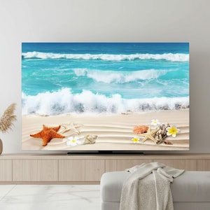 Op de afbeelding: Een grote, rechthoekige wandkunstprint met een strandscène. Het kunstwerk toont een turquoise oceaan met witte golven, een zandstrand en diverse schelpen, zeesterren en bloemen. De print wordt weergegeven op een licht houten kast.