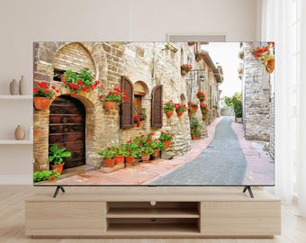 May include: A large flat-screen television displays a scenic image of a European street lined with buildings, doors, and windows. The buildings are made of stone and brick, with red flowers in pots. The TV is on a light wood cabinet.