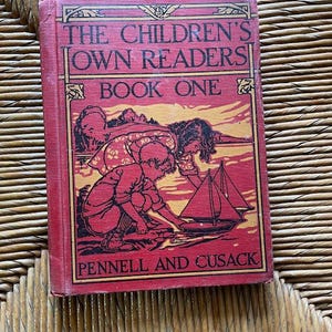 Pode incluir: Um livro vermelho vintage intitulado "THE CHILDREN'S OWN READERS BOOK ONE" com uma ilustração de crianças e um veleiro. A capa tem uma borda decorativa e os nomes dos autores, PENNELL AND CUSACK, estão impressos na parte inferior.