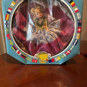 Puede incluir: Una muñeca vintage en una caja. La muñeca tiene cabello rubio y lleva un vestido largo con un estampado floral. La caja es azul claro y tiene una ventana circular con banderas alrededor del borde. La muñeca está sentada.
