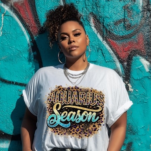 Op de afbeelding: Wit T-shirt met de tekst "AQUARIUS Season" in een luipaardprint met gouden en turquoise accenten. Het shirt wordt gedragen door een persoon voor een met graffiti bedekte muur.
