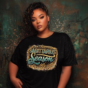 Op de afbeelding: Zwart T-shirt met de tekst "SAGITTARIUS Season" in goud, turquoise en luipaardprint. Het shirt wordt gedragen door een persoon met krullend haar en oorringen. De achtergrond heeft een turquoise en goudkleurig kleurenschema.