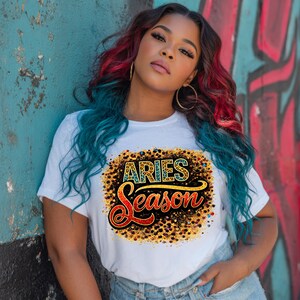 Op de afbeelding: Wit T-shirt met de tekst "ARIES Season" in een kleurrijk ontwerp. Het shirt heeft een ronde hals en korte mouwen. Het ontwerp bevat goud, rood en zwart. De persoon op de foto draagt een blauwe spijkerbroek.