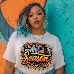 Op de afbeelding: Wit T-shirt met de tekst "CANCER Season" in een kleurrijk luipaardprint ontwerp. Het shirt heeft een ronde hals en korte mouwen. De achtergrond is een kleurrijke graffiti muur.