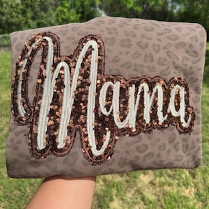 May include: A taupe-colored sweatshirt with a leopard print pattern. The word "Mama" is embroidered on the front in a cursive font, outlined in white and filled with copper-colored sequins. The sweatshirt is held up against a green background.