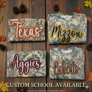 May include: Four camouflage sweatshirts with school names embroidered in glittery lettering: Texas, Mizzou, Aggies, and Bobcats. The sweatshirts are displayed on a wooden surface with fall-themed decorations. Text at the bottom reads "CUSTOM SCHOOL AVAILABLE."