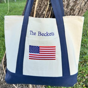 May include: Cream-colored tote bag with navy blue handles and base. The front pocket features an embroidered American flag and the text "The Becketts" in navy blue.