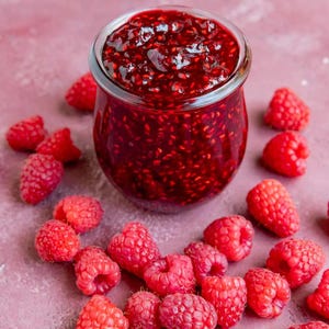 May include: A jar of homemade raspberry jam with fresh raspberries scattered around it. The jam is a deep red color and has a thick, glossy texture. The raspberries are bright red and plump, with a slightly textured surface. The background is a solid pink color.