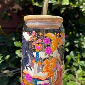 May include: A clear glass tumbler with a bamboo lid and a glass straw. The tumbler is decorated with a fantasy-themed design featuring dragons, a sword, books, a butterfly, and roses. The design includes orange, pink, and black elements.