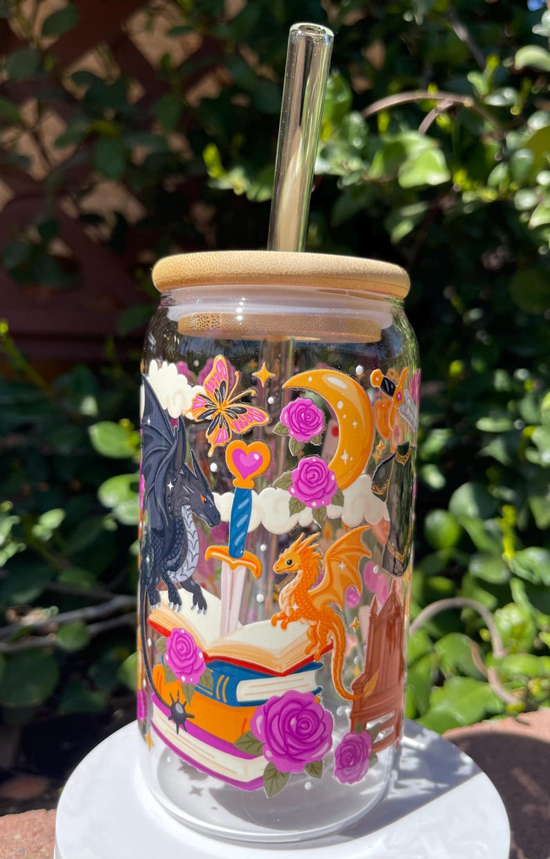 May include: A clear glass tumbler with a bamboo lid and a glass straw. The tumbler is decorated with a fantasy-themed design featuring dragons, a sword, books, a butterfly, and roses. The design includes orange, pink, and black elements.
