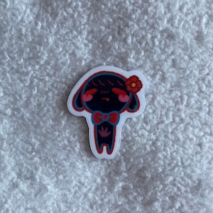 May include: A die-cut sticker featuring a cartoon character with a black body, red accents, and a red bow tie. The character has a red flower on its head and pink cheeks. The sticker is on a white background.