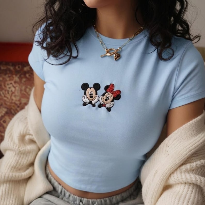 May include: A light blue short-sleeved t-shirt featuring an embroidered graphic of Mickey Mouse and Minnie Mouse on the chest. The characters are depicted with their hands resting under their chins. The shirt is worn with a gold-toned chain necklace.
