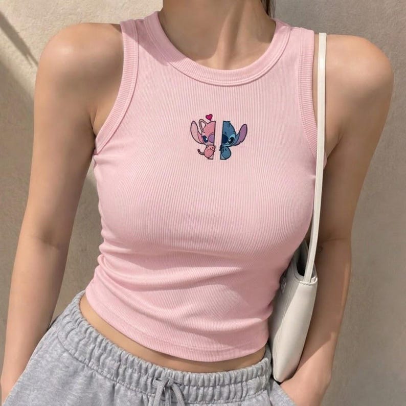 May include: A light pink ribbed tank top featuring a small embroidered graphic on the chest. The design shows two split halves of a blue and pink alien character facing each other, with a small red heart positioned above the center gap.