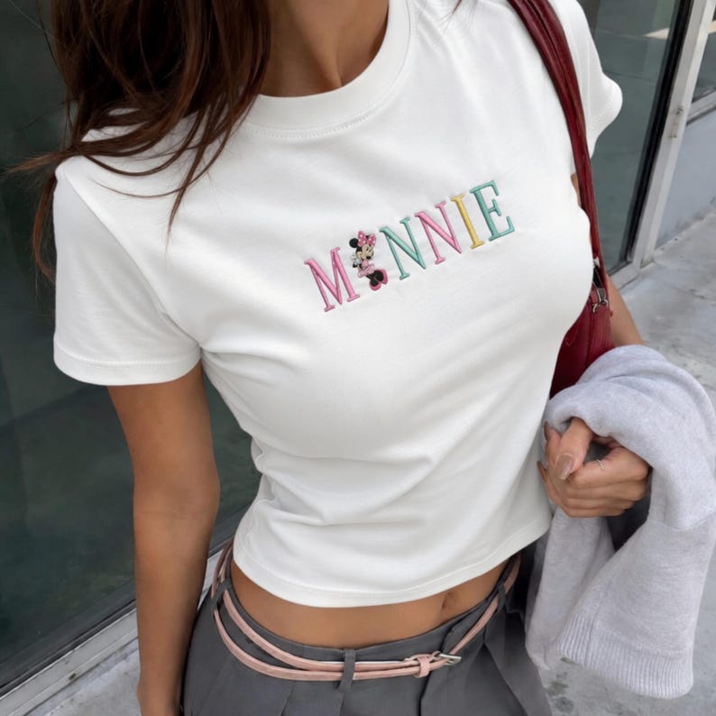Puede incluir: Camiseta blanca con la palabra "MINNIE" bordada en colores pastel. La letra "O" est&aacute; reemplazada por un gr&aacute;fico de Minnie Mouse. La camiseta tiene mangas cortas y cuello redondo.