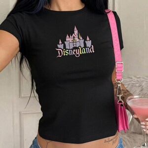 May include: Black short-sleeved t-shirt featuring a Disneyland castle graphic in pink, purple, and grey. The word "Disneyland" is printed below the castle in a cursive font. The shirt has a fitted style.