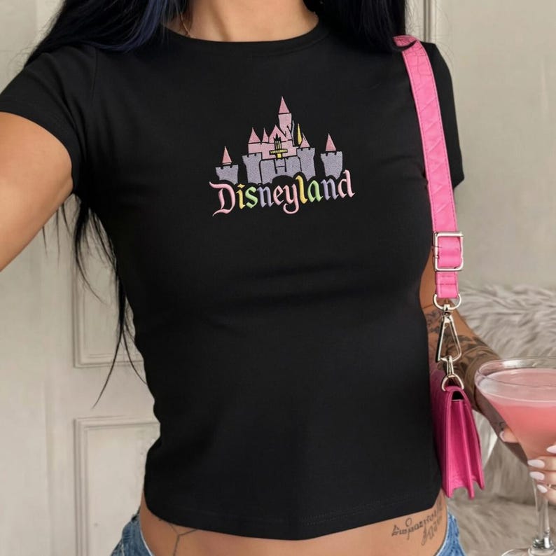May include: Black short-sleeved t-shirt featuring a Disneyland castle graphic in pink, purple, and grey. The word "Disneyland" is printed below the castle in a cursive font. The shirt has a fitted style.