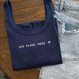 Puede incluir: Camiseta sin mangas acanalada azul marino con el texto "AIR PLANE MODE" y un peque&ntilde;o gr&aacute;fico de avi&oacute;n blanco. Se combina con unos shorts vaqueros de lavado claro, creando un conjunto informal. La camiseta est&aacute; doblada sobre una superficie de madera.