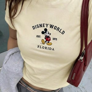 May include: A cream-coloured t-shirt with the Disney World logo, featuring Mickey Mouse. The logo includes the text "DISNEY WORLD", "EST. 1971", and "FLORIDA". The shirt has a round neckline and short sleeves.