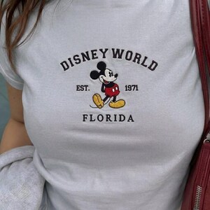 May include: Pale blue t-shirt featuring a Mickey Mouse graphic and the text "DISNEY WORLD FLORIDA". The graphic includes the text "EST. 1971". The shirt is a crew neck style.