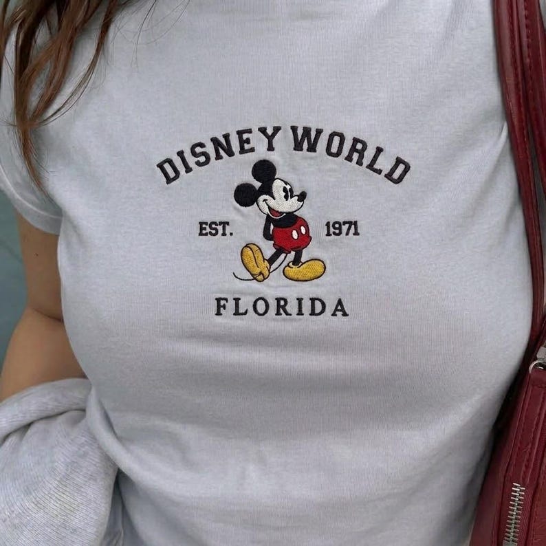 May include: Pale blue t-shirt featuring a Mickey Mouse graphic and the text "DISNEY WORLD FLORIDA". The graphic includes the text "EST. 1971". The shirt is a crew neck style.
