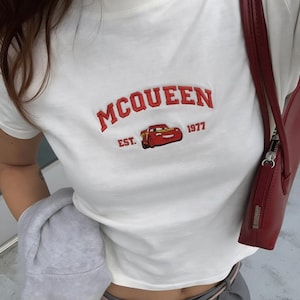 以下が含まれることがあります： 赤い文字で「MCQUEEN」と刺繍された白いTシャツ。赤い漫画の車のグラフィックと「EST. 1977」の文字が特徴です。画像の右側には赤いハンドバッグが見えます。