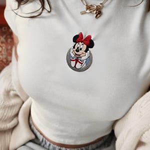 May include: White short-sleeved shirt with a Minnie Mouse patch. The patch features Minnie Mouse with a red bow, white gloves, and red and white shoes. A gold necklace with a charm is also visible.