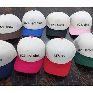 Puede incluir: Una colecci&oacute;n de gorras de b&eacute;isbol con coronas blancas y viseras de colores. Las gorras son de varios colores, incluyendo marr&oacute;n, azul claro, negro, rosa, azul marino, rosa intenso, rojo y verde bosque. Cada gorra tiene una etiqueta numerada.