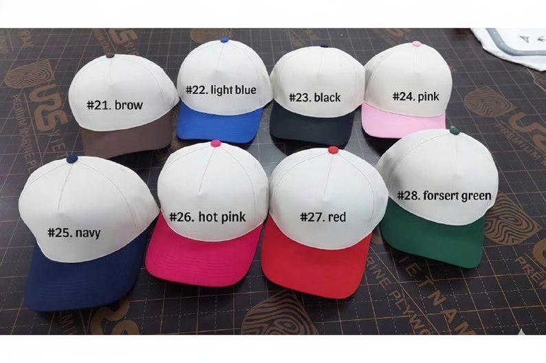 Puede incluir: Una colecci&oacute;n de gorras de b&eacute;isbol con coronas blancas y viseras de colores. Las gorras son de varios colores, incluyendo marr&oacute;n, azul claro, negro, rosa, azul marino, rosa intenso, rojo y verde bosque. Cada gorra tiene una etiqueta numerada.