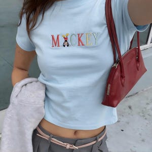 May include: Pale blue t-shirt featuring the embroidered word "MICKEY" in various colours, with a cartoon character. A red handbag with a zip is visible. The outfit includes grey trousers and a pink belt.