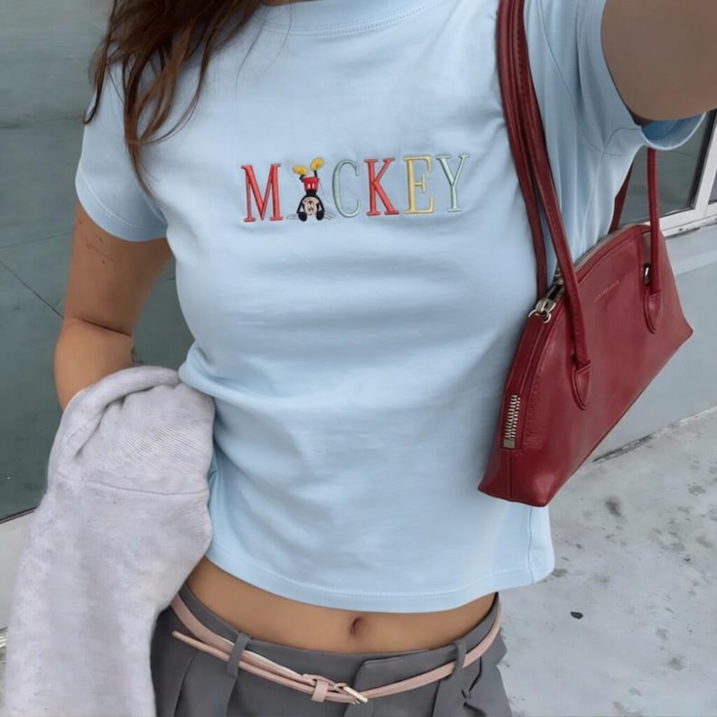 May include: Pale blue t-shirt featuring the embroidered word "MICKEY" in various colours, with a cartoon character. A red handbag with a zip is visible. The outfit includes grey trousers and a pink belt.