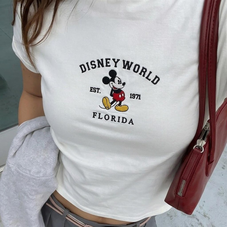 May include: White t-shirt with an embroidered Mickey Mouse design and the text "DISNEY WORLD FLORIDA EST. 1971". The top has a crew neck and short sleeves. A red handbag is visible on the right.