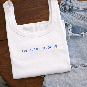 Puede incluir: Camiseta sin mangas blanca con costuras acanaladas y la frase azul "AIR PLANE MODE", acompa&ntilde;ada de un peque&ntilde;o avi&oacute;n. Se combina con unos shorts vaqueros de lavado claro, creando un look casual y viajero.