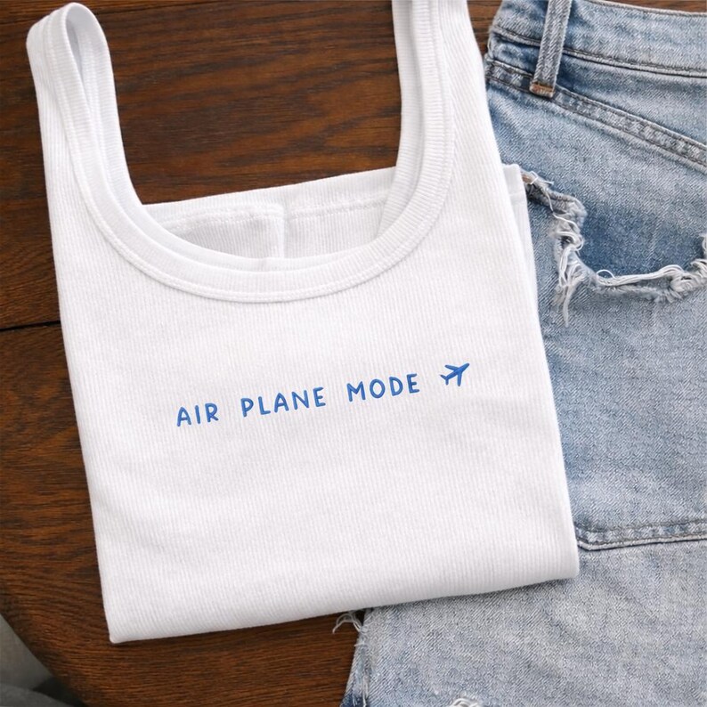 Puede incluir: Camiseta sin mangas blanca con costuras acanaladas y la frase azul "AIR PLANE MODE", acompa&ntilde;ada de un peque&ntilde;o avi&oacute;n. Se combina con unos shorts vaqueros de lavado claro, creando un look casual y viajero.