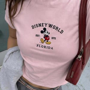 May include: A pale pink t-shirt with the Disney World logo and Mickey Mouse. The text reads "DISNEY WORLD EST. 1971 FLORIDA". The shirt has a crew neck and short sleeves.