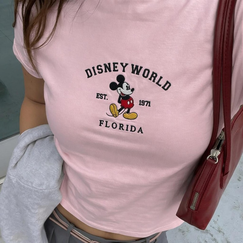 May include: A pale pink t-shirt with the Disney World logo and Mickey Mouse. The text reads "DISNEY WORLD EST. 1971 FLORIDA". The shirt has a crew neck and short sleeves.