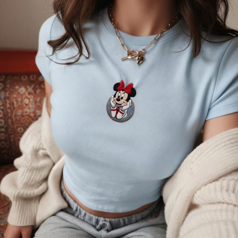 May include: Light blue t-shirt featuring an embroidered Minnie Mouse design. The shirt has short sleeves and a round neckline. A gold-colored necklace with a heart pendant is also visible.