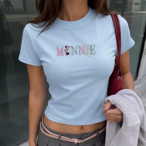 Puede incluir: Camiseta azul claro con la palabra "MINNIE" bordada en letras coloridas. La camiseta tiene mangas cortas y cuello redondo. Un bolso rojo es visible en el fondo.