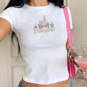 May include: White t-shirt featuring a pastel-coloured Disneyland castle graphic and the word "Disneyland" below. The shirt has short sleeves and a fitted shape. A pink handbag is visible on the right.