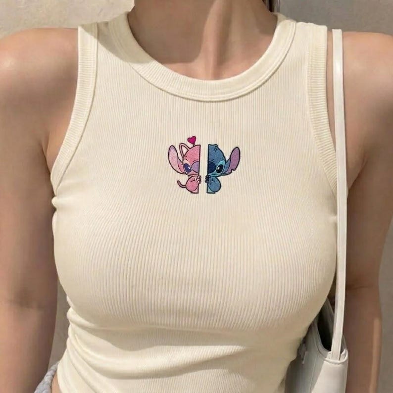 May include: A cream-colored ribbed tank top featuring a central embroidered graphic of two split characters. The left side is a pink creature with a small heart, and the right side is a blue creature, both facing inward toward the center seam.