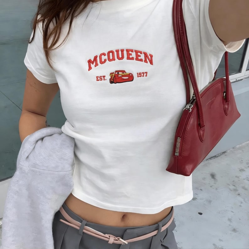 以下が含まれることがあります： 赤い文字で「MCQUEEN EST. 1977」と書かれた白いTシャツ。赤い漫画の車のグラフィックが特徴です。ジッパー付きの赤いハンドバッグが見えます。シャツはグレーのパンツとピンクのベルトと組み合わせています。