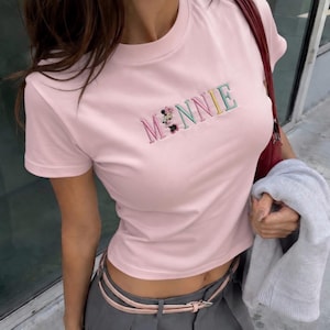 Puede incluir: Una camiseta rosa claro con la palabra "MINNIE" bordada en letras coloridas. La letra "M" presenta un dise&ntilde;o de Minnie Mouse. La camiseta tiene mangas cortas y cuello redondo.